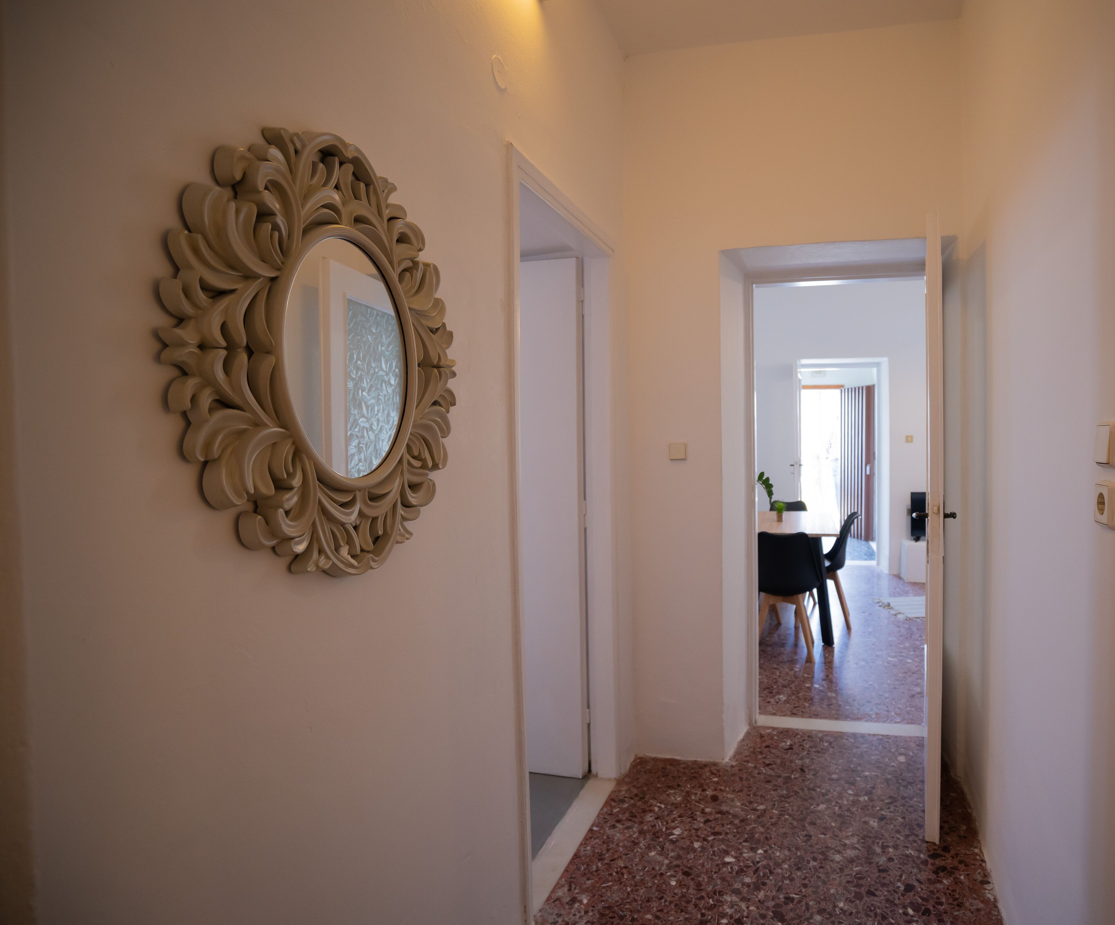 A hallway with a decorative mirror on the left, a door leading to a dining area, and a tiled floor.