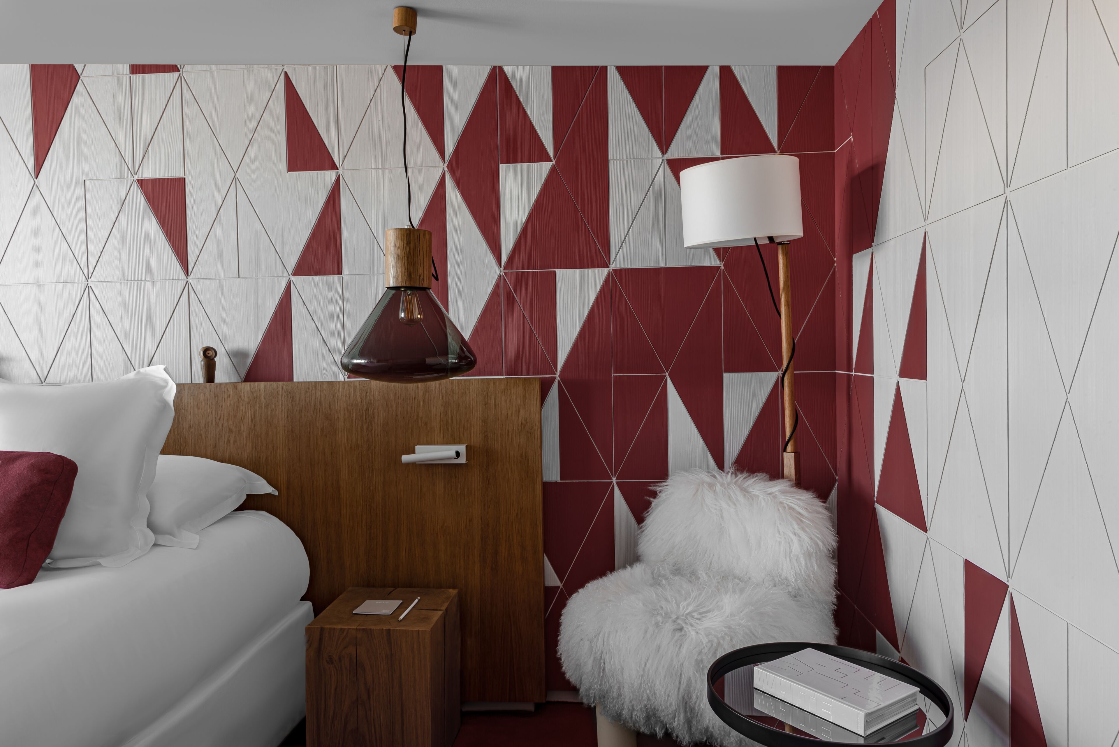 Una habitación de hotel moderna con una pared de diseño geométrico, un armario de madera, un sillón blanco mullido, una mesita de noche con lámpara y una cama con ropa de cama blanca y una almohada roja.