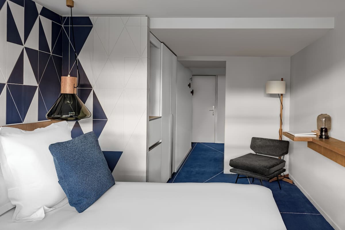 A hotel room with a bed, a chair, a desk with a lamp, and a geometric-patterned wall.