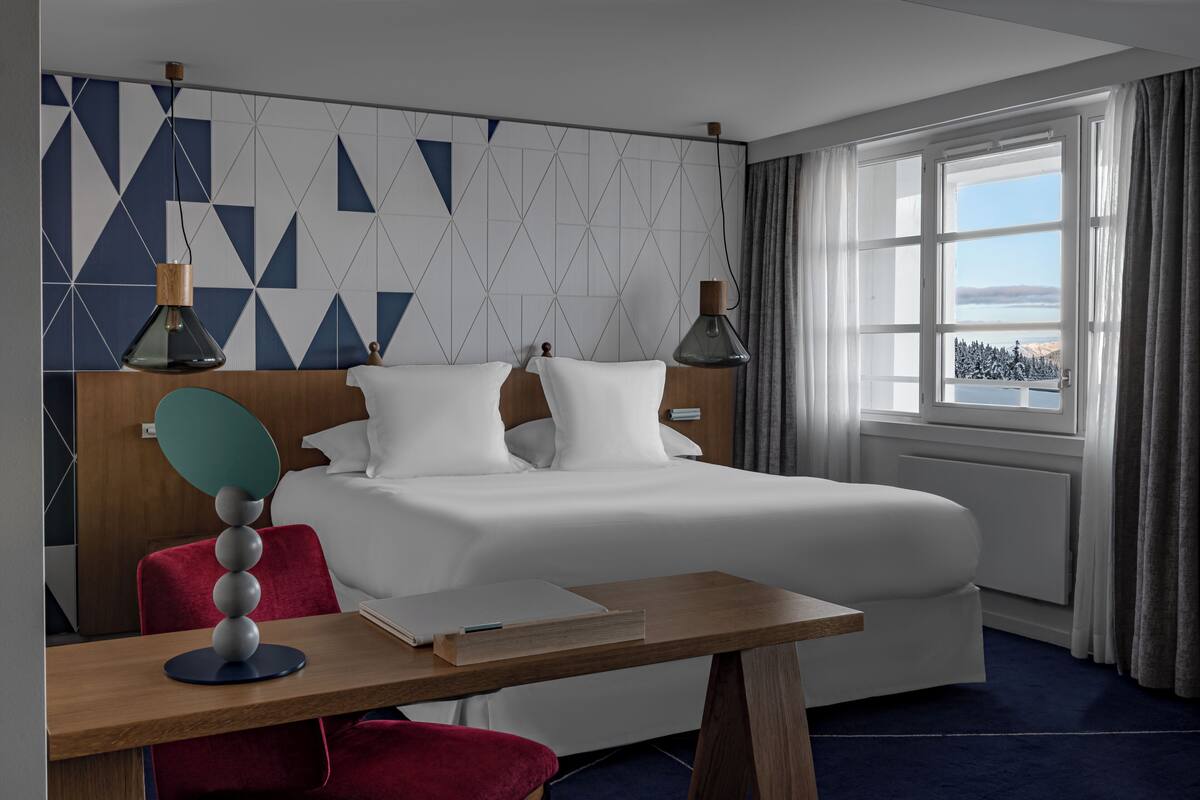 A modern hotel room with a large bed, a desk with a notebook, a red chair, and a view of a snowy landscape through a window.