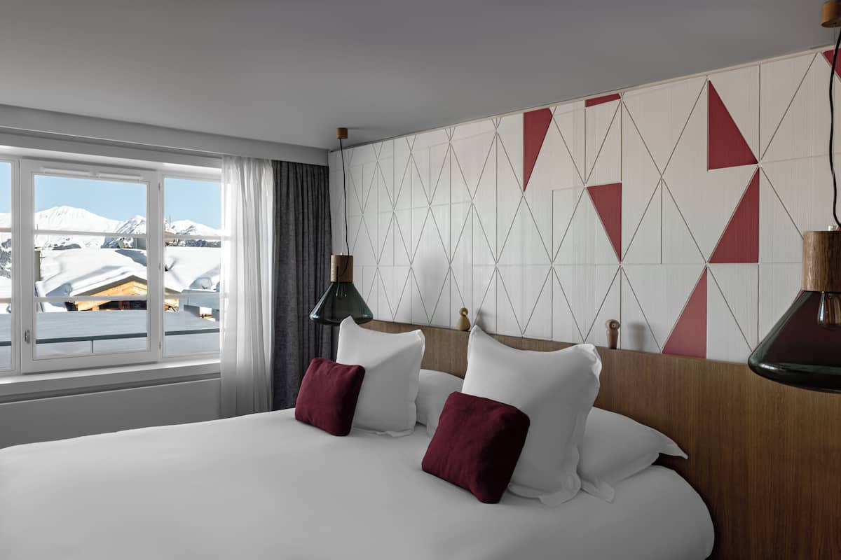 A hotel room with a large bed, two red pillows, a wooden headboard, and a view of snow-capped mountains through a window.