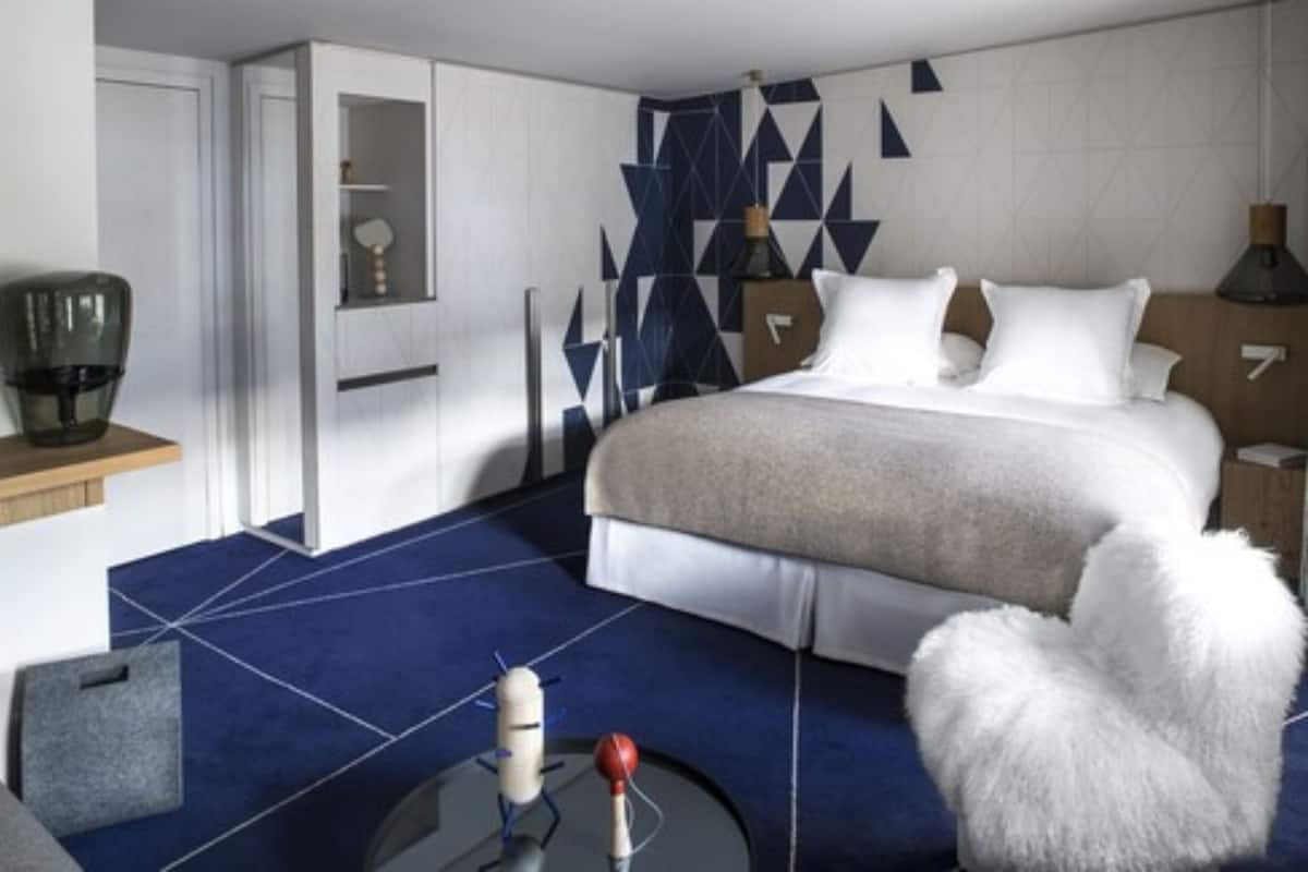A modern hotel room with a large bed, a white furry chair, a coffee machine, and a geometric-patterned wall.