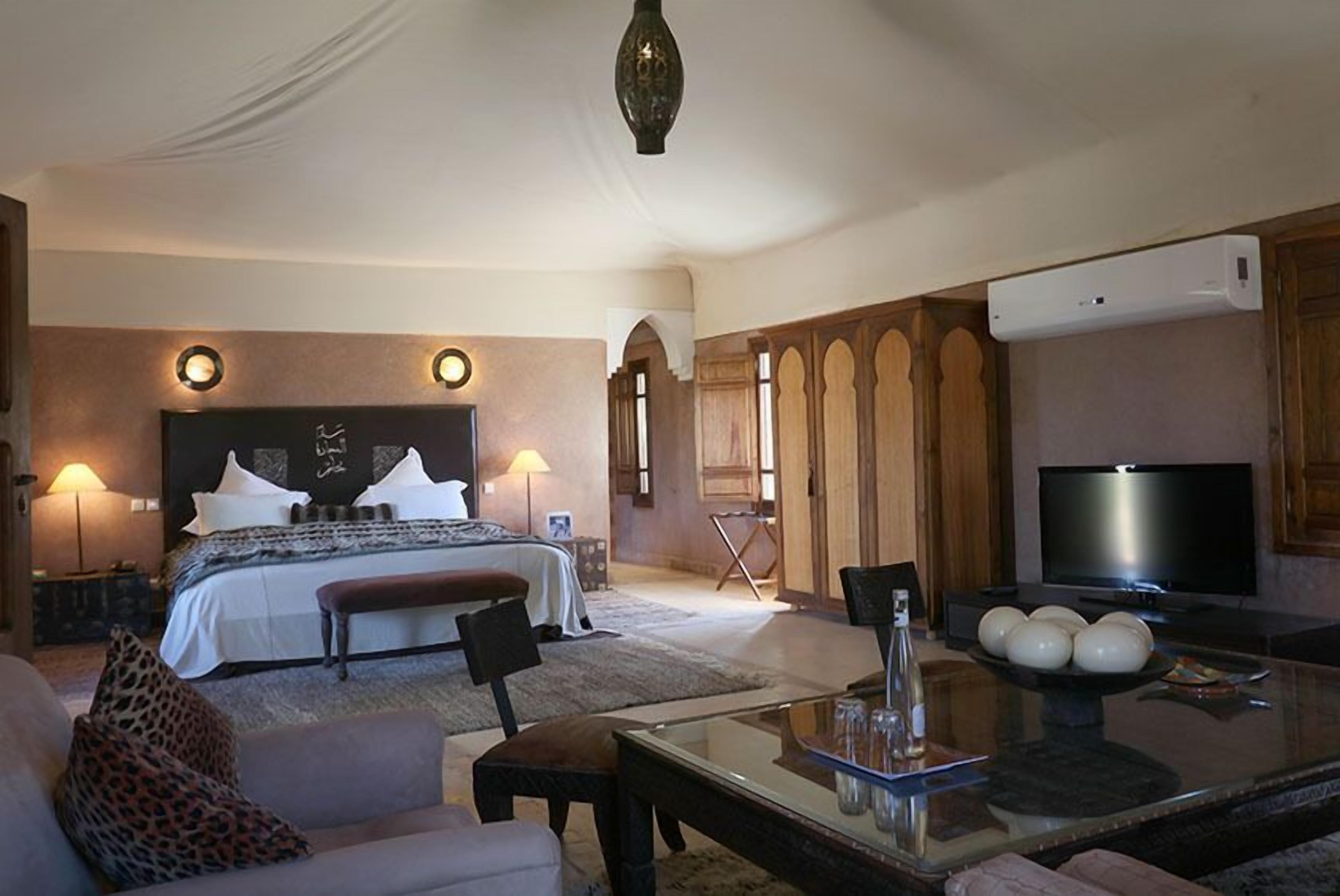 A hotel room with a large bed, a TV, a glass coffee table, and a seating area with a sofa and chairs.