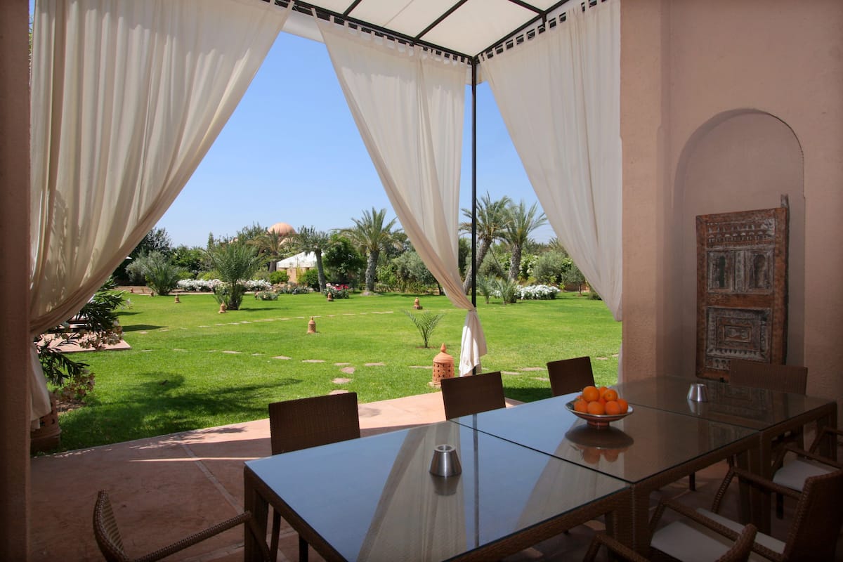 A dining area with a glass table, white curtains, and a view of a garden with palm trees and a building in the background.