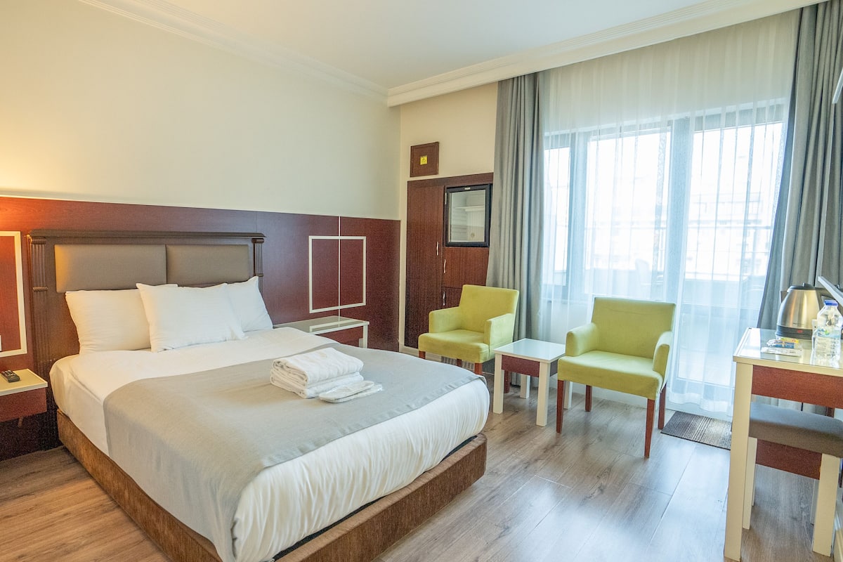A hotel room with a large bed, bedside tables, a desk with a chair, a window with curtains, and a wall-mounted TV.