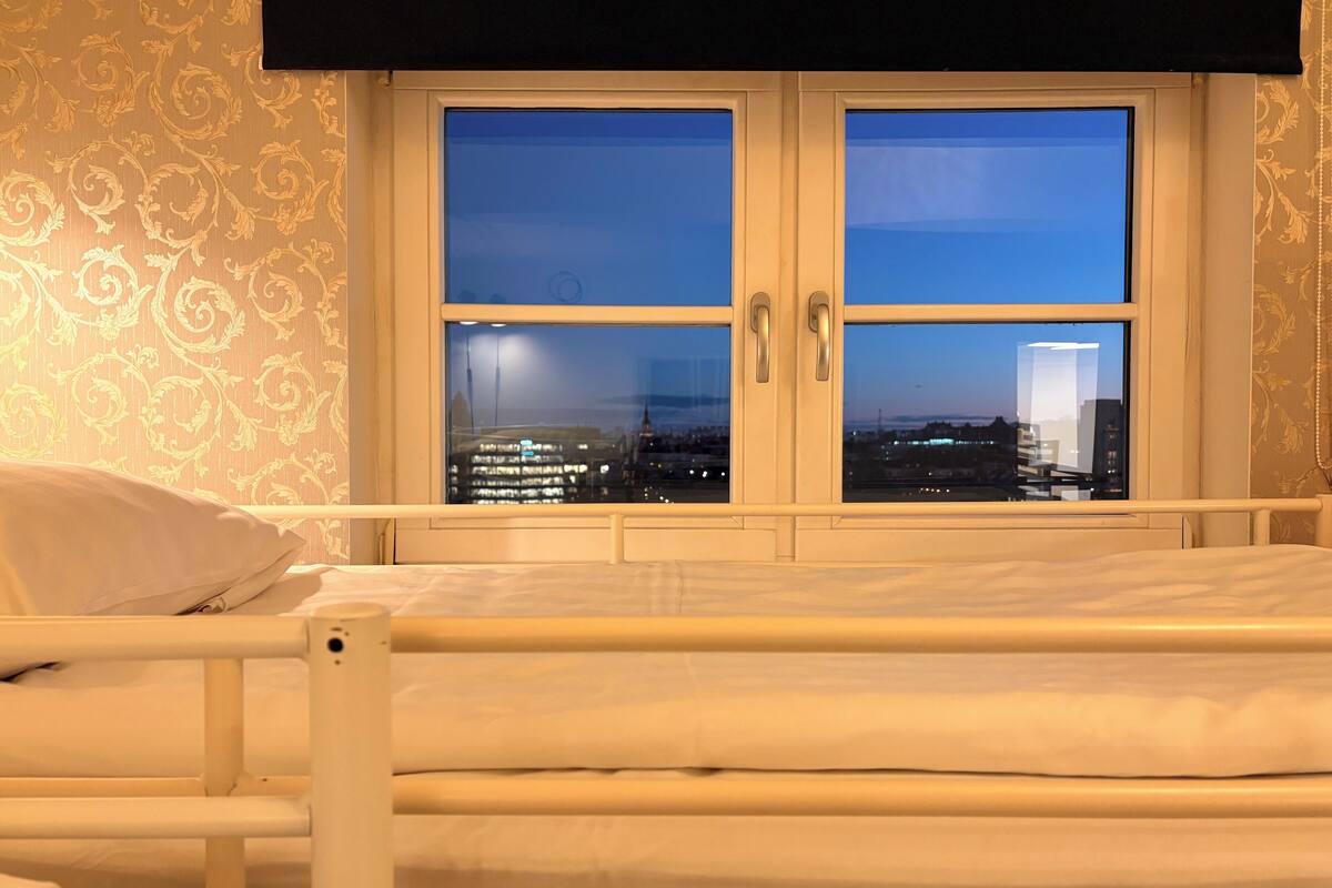 A bedroom with a bed, a window with two doors, and a view of a cityscape at dusk.