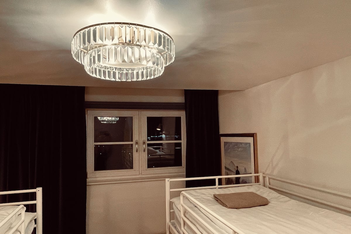 A bedroom with a bed, a window, a chandelier, and a framed picture on the wall.