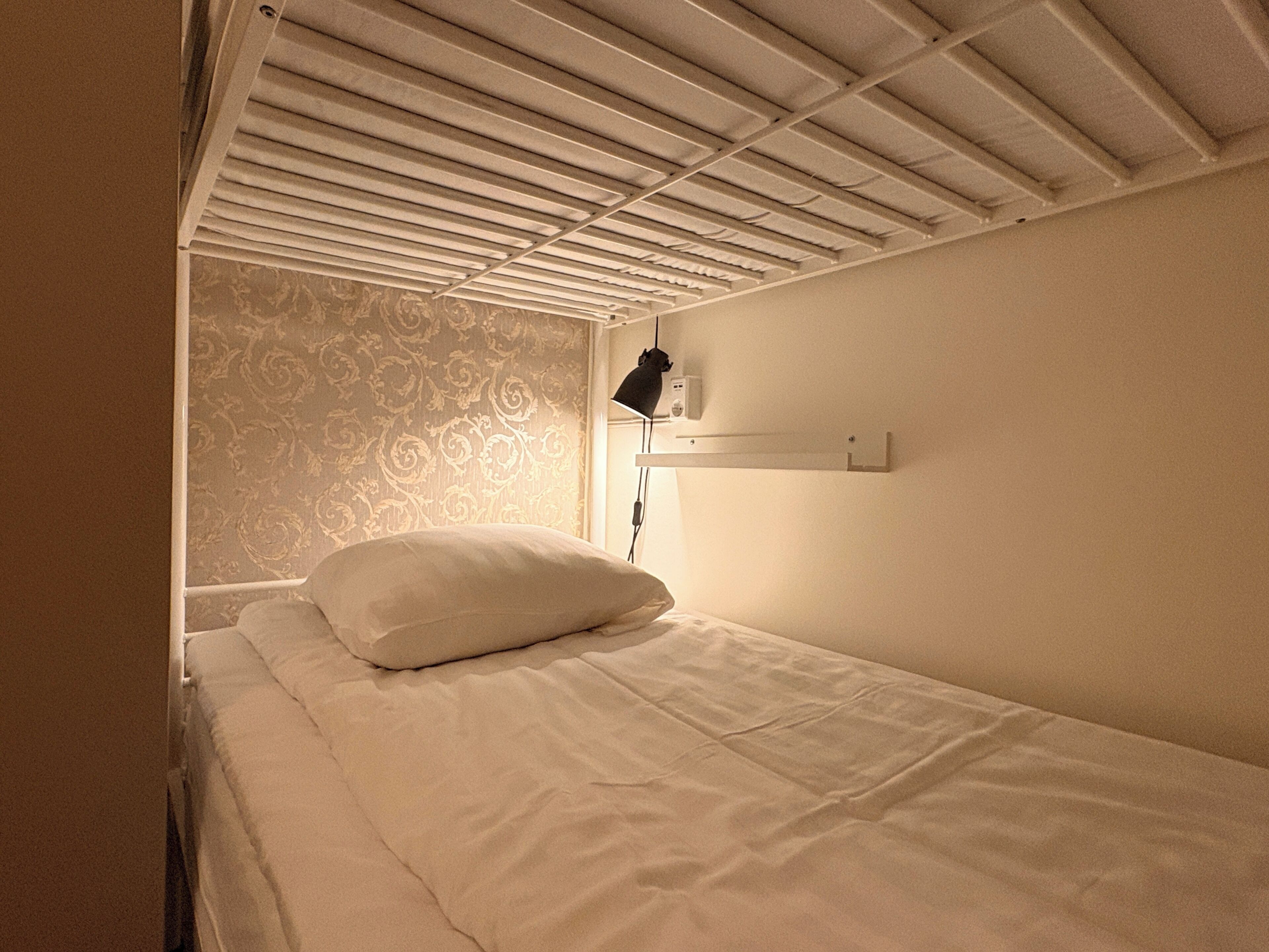 A single bed with a white pillow, a black lamp on a bedside table, and a patterned wallpaper on the wall.