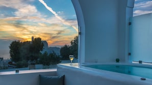 Un espacio exterior moderno con una mesa de vidrio y una copa de vino blanco, con vistas a un paisaje urbano y un atardecer.