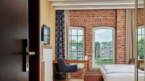 A hotel room with a brick wall, a desk with a chair, a bed, and large windows offering a view of buildings and greenery. - Hotel Hafenspeicher (Leer)