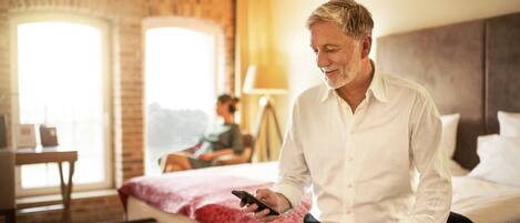 Un hombre sentado en una cama en una habitación de hotel, usando un teléfono inteligente. La habitación tiene una pared de ladrillo, una ventana y una lámpara.