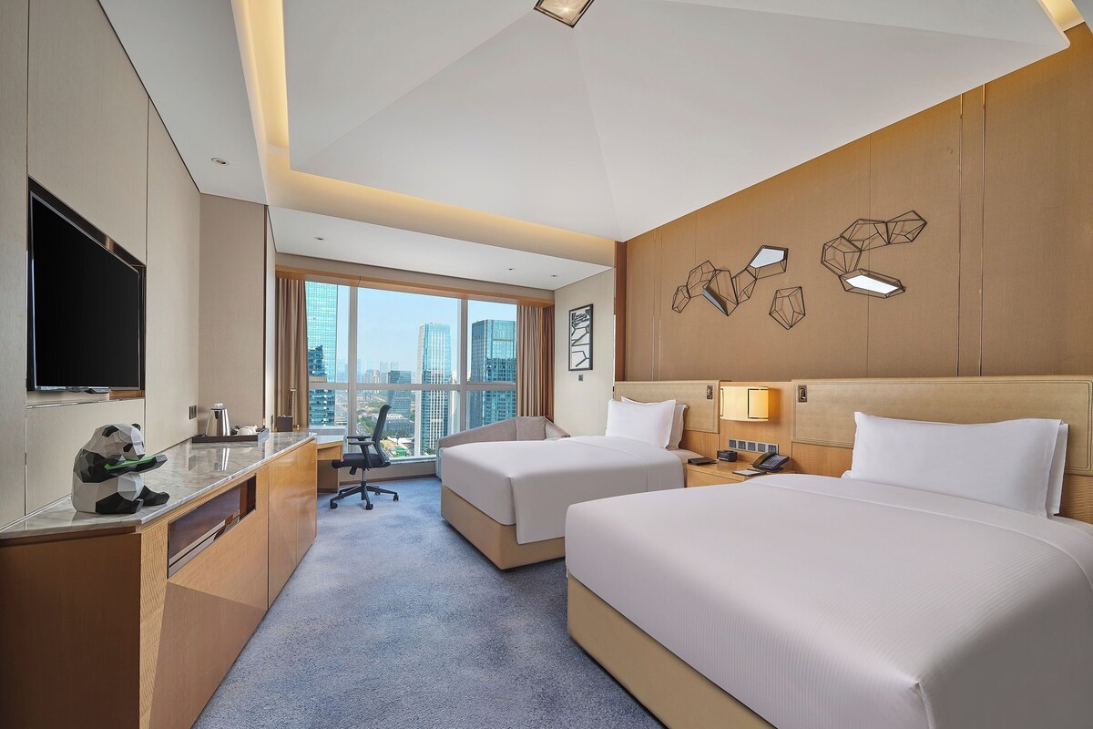 A hotel room with two beds, a desk with a flat-screen TV, a coffee maker, and a view of the cityscape.