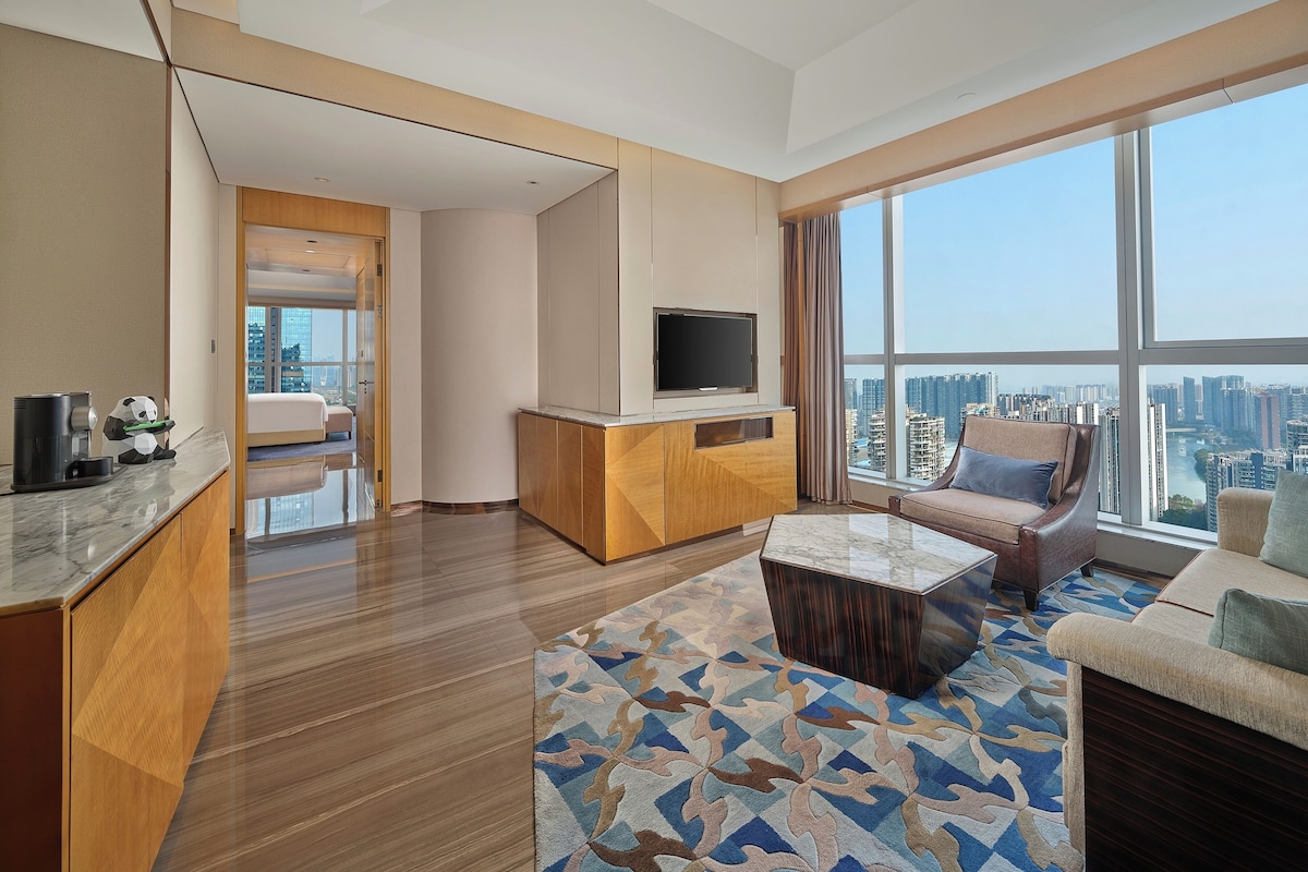 A modern hotel room with a large window, a TV on a wooden cabinet, a sofa, and a coffee table.