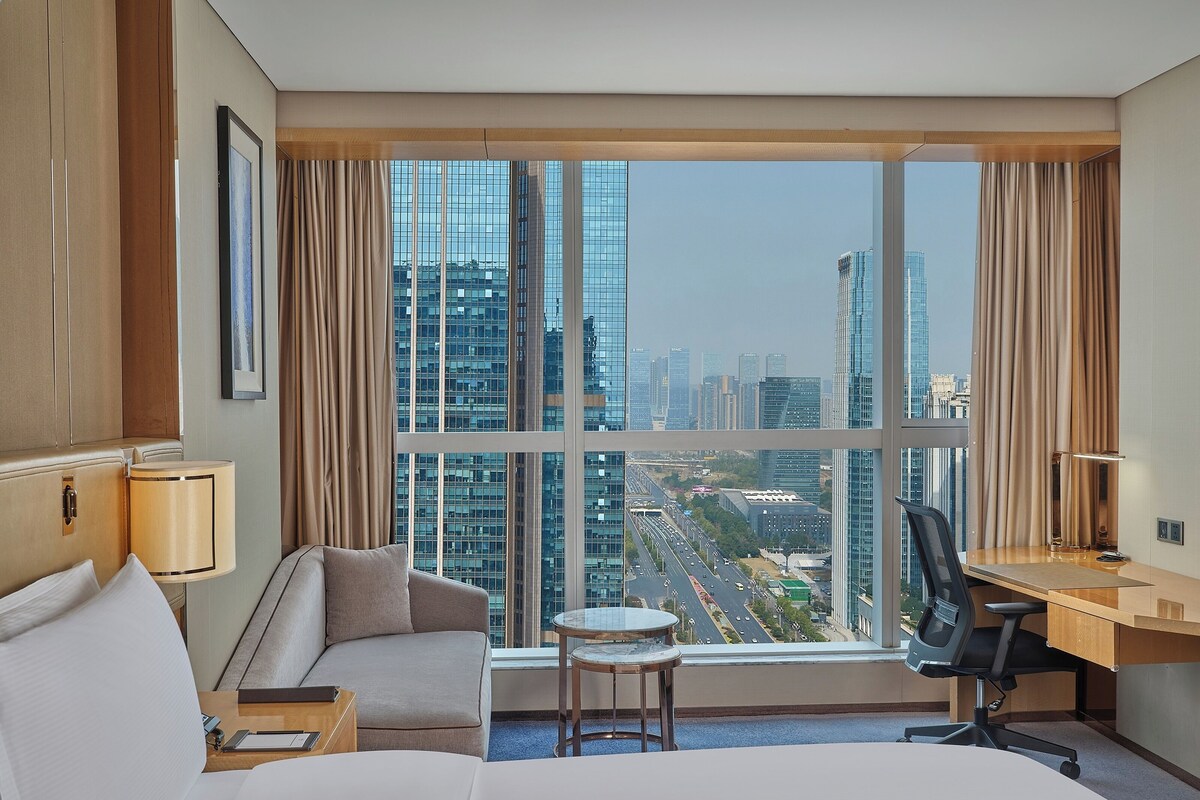 A hotel room with a bed, a desk, a chair, a small table, and a view of a cityscape through large windows.