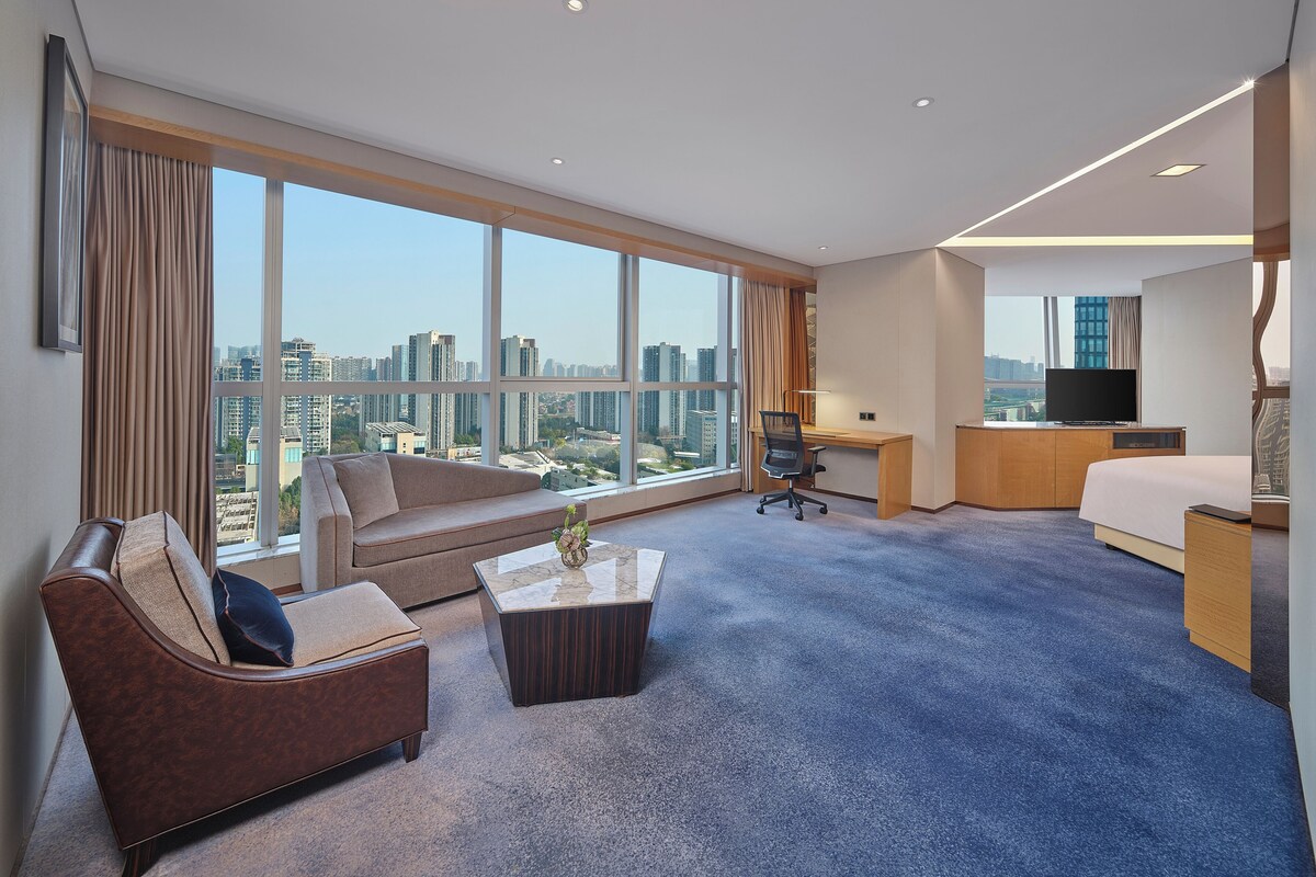 A modern hotel room with a large window, a sofa, a desk, a bed, and a television.