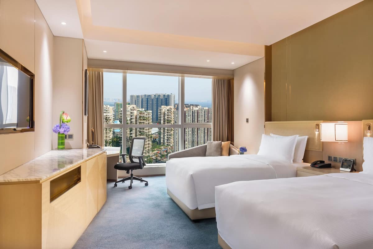 A hotel room with two beds, a desk, and a large window offering a city view.