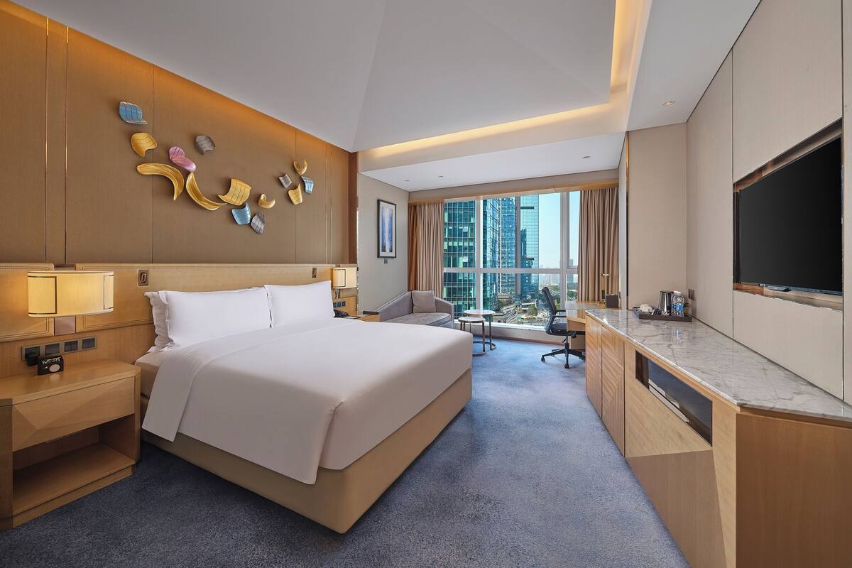 A hotel room with a large bed, a desk with a flat-screen TV, a desk lamp, a chair, and a view of the cityscape.