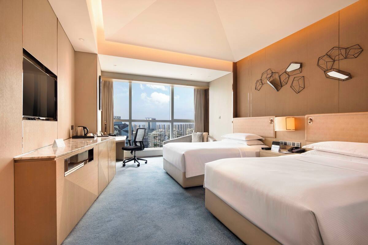A hotel room with two beds, a desk, a TV, and a city view.
