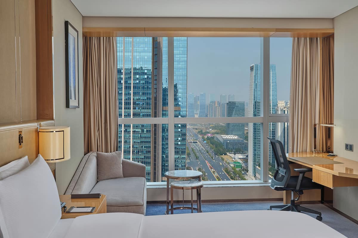 A hotel room with a bed, a desk, a chair, a small table, and a view of a cityscape through large windows.