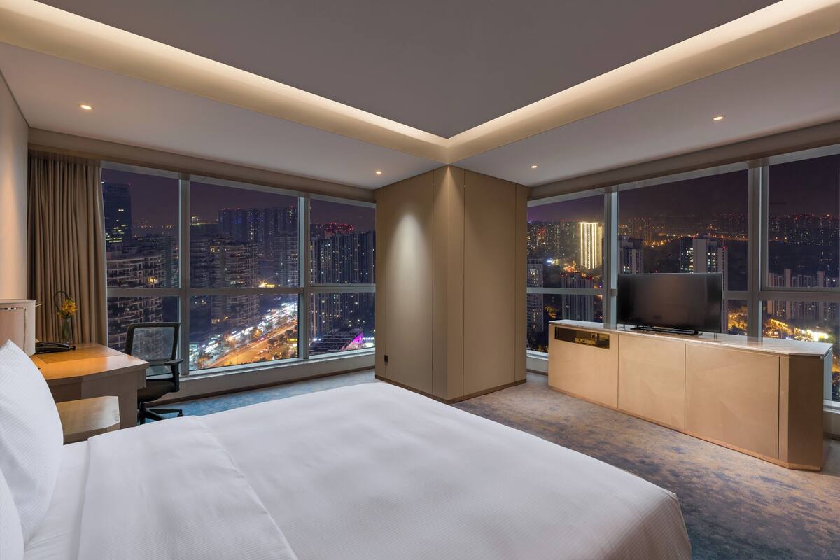 A hotel room with a large bed, a desk, a TV, and a view of a cityscape at night.