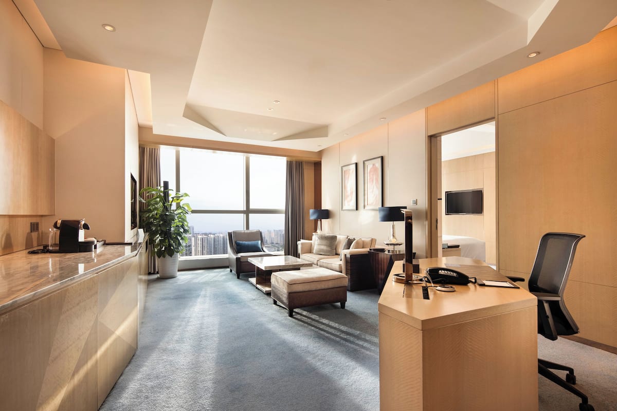 A modern hotel room with a kitchen area, a seating area with a sofa and ottoman, a desk with a telephone and lamp, and a large window offering a city view.