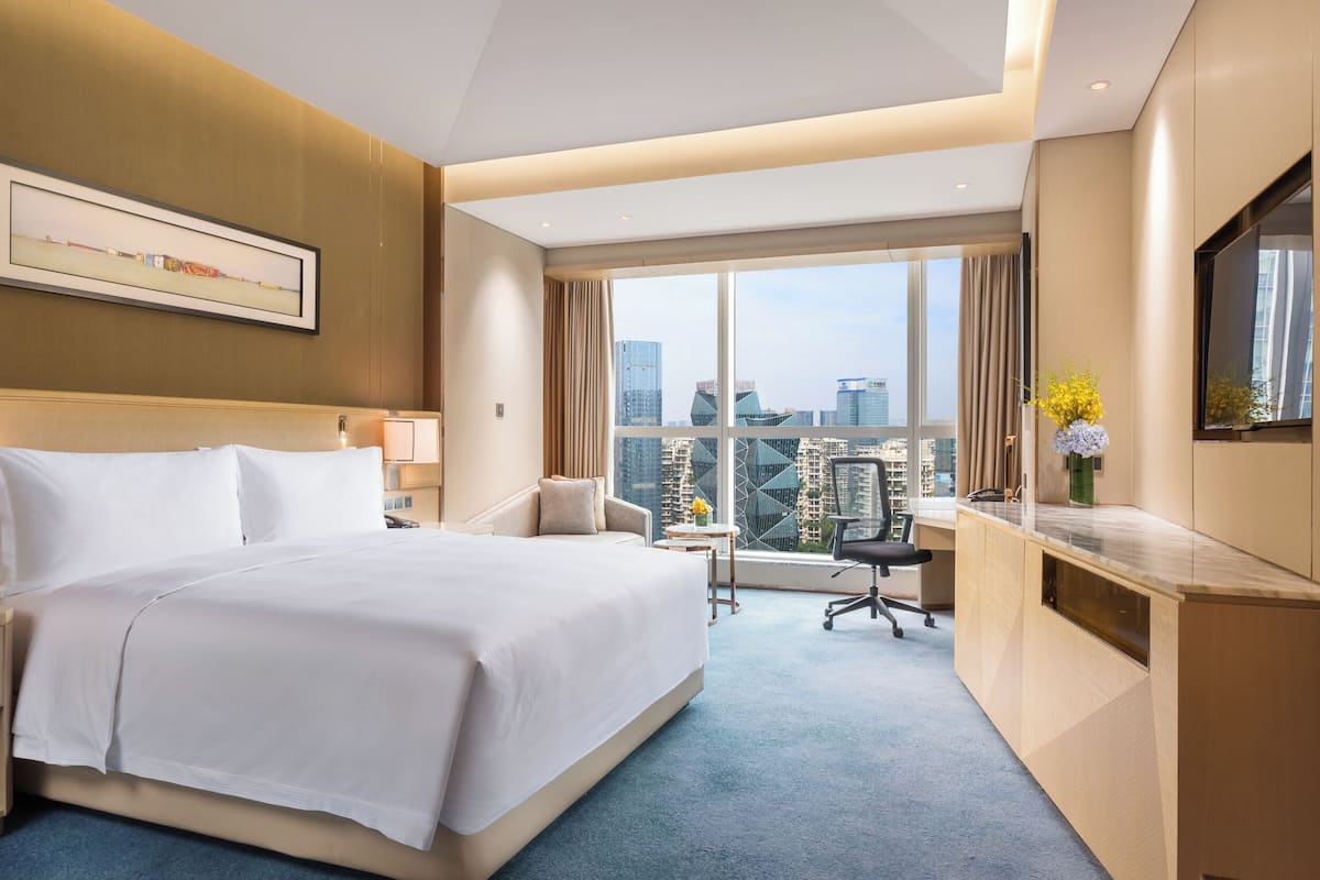 A hotel room with a large bed, a desk, a chair, a TV, and a window with a city view.