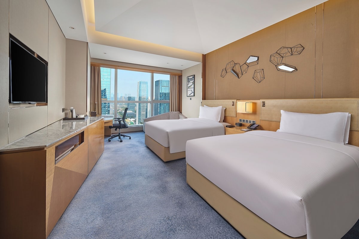 A hotel room with two beds, a desk, a TV, and a city view.