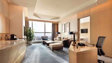 Una habitación de hotel moderna con un ventanal grande, una zona de estar con sofá y sillones, un escritorio con teléfono y una cafetera.