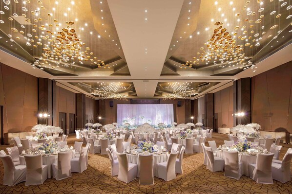 A banquet hall with round tables, white chairs, and chandeliers.