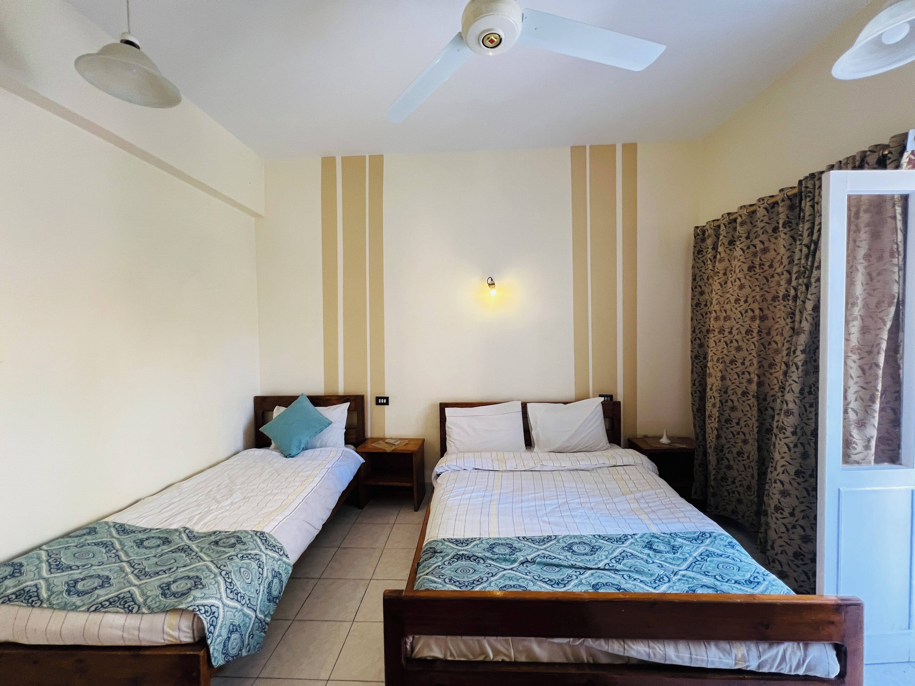 Una habitación con dos camas individuales, ventilador de techo, luz empotrada en la pared y ventana con cortinas.