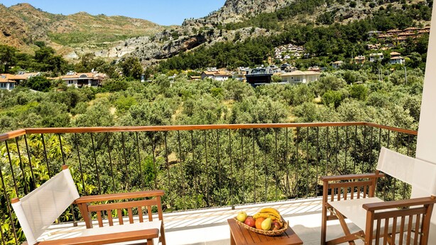 A balcony with wooden furniture and a fruit basket, overlooking a hillside with houses. - Kerem Apart Hotel Gocek (Fethiye)