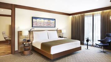 A hotel room with a large bed, a bedside table with a lamp, a chair, a small table with a plant, and a painting on the wall.