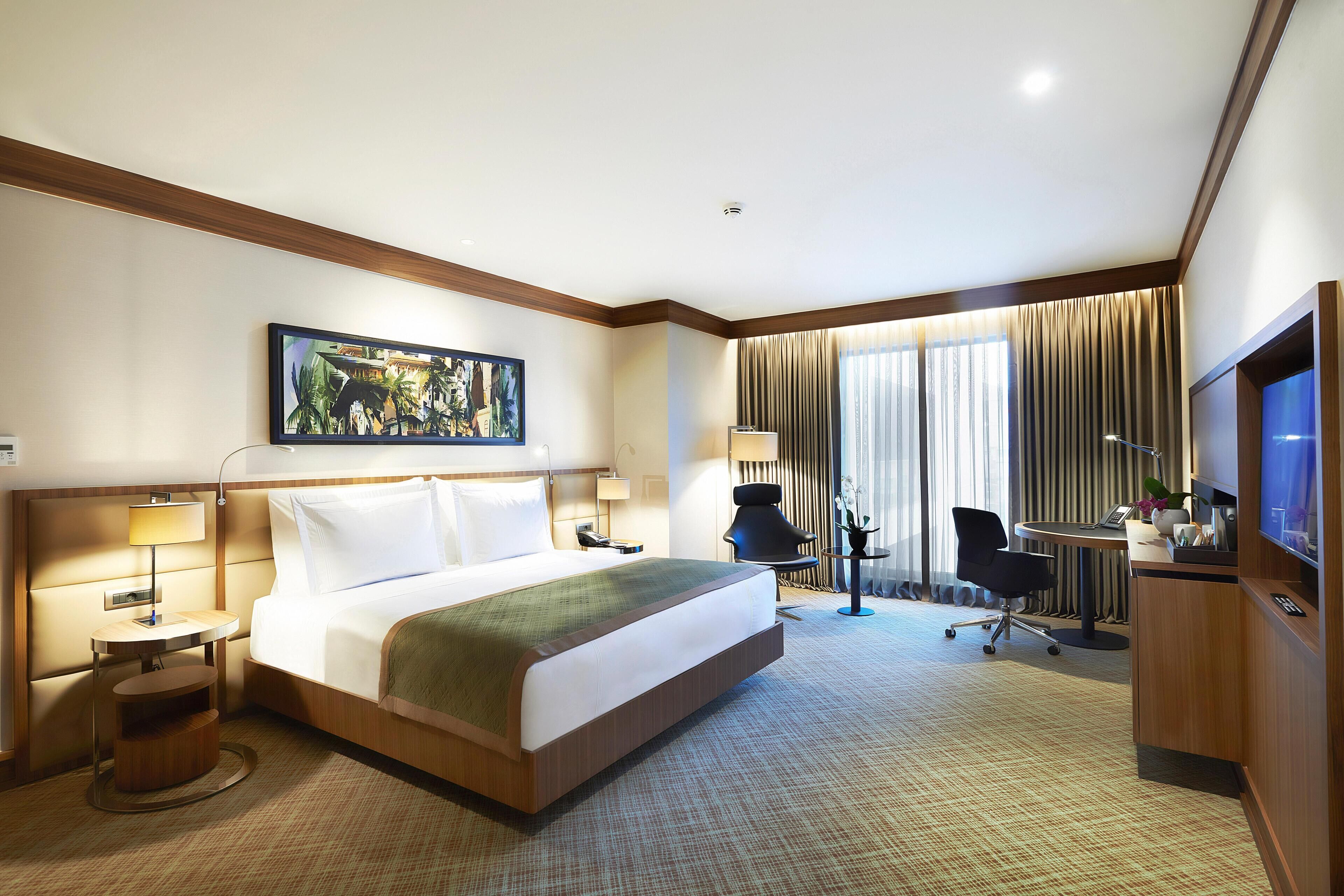 A hotel room with a bed, bedside tables, a desk, a chair, a TV, and a large window with curtains.