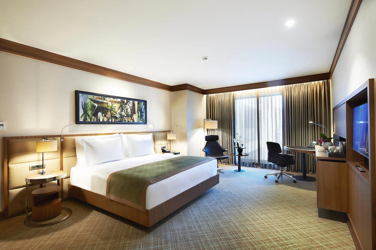 A hotel room with a bed, bedside tables, a desk, a chair, a TV, and a large window with curtains.
