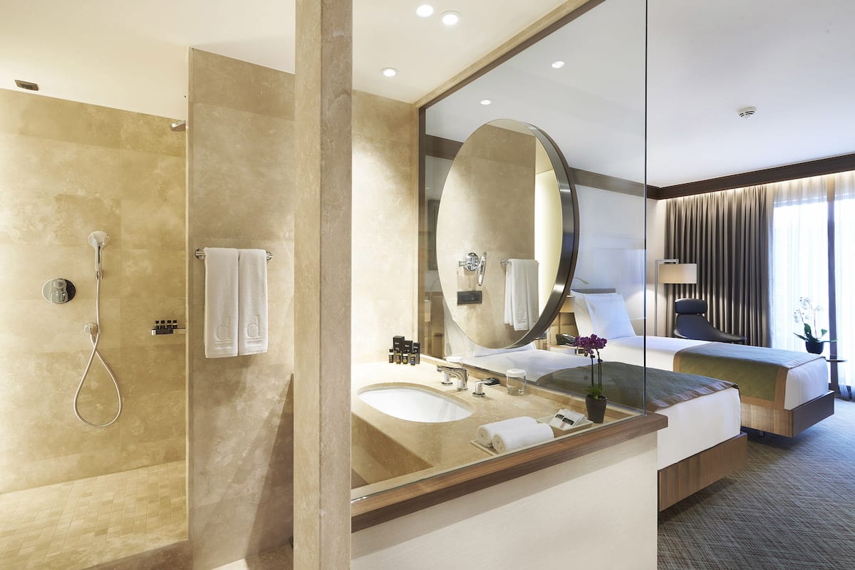 A modern hotel bathroom with a glass-enclosed shower, a large mirror, a sink, and a bed with a view of the outdoors.