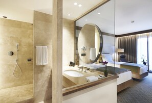 A modern hotel bathroom with a glass-enclosed shower, a large mirror, a sink, and a bed with a view of the outdoors. - Divan Adana (Adana)