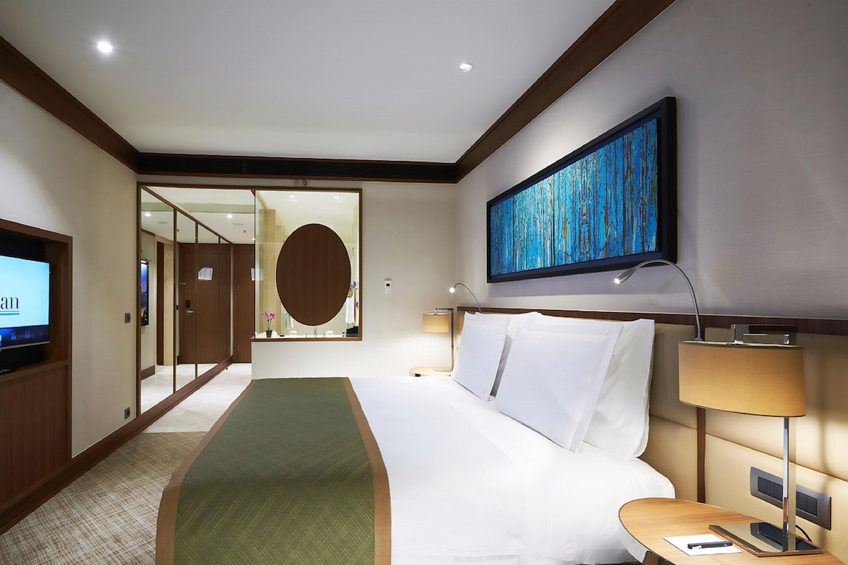 A hotel room with a bed, a TV, a desk, a lamp, a painting, and a mirror.