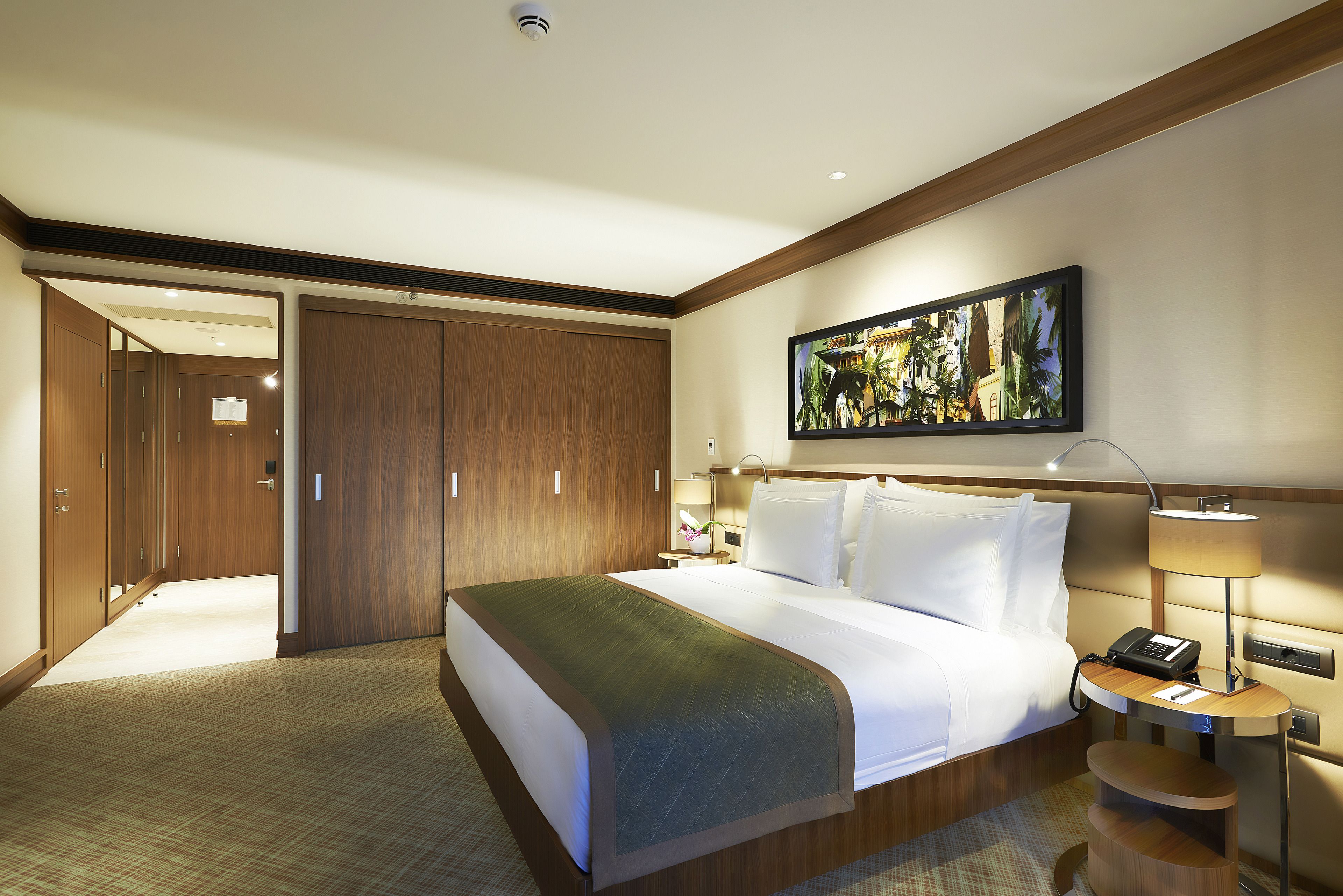 A hotel room with a large bed, a desk with a lamp, a painting on the wall, and a wardrobe.