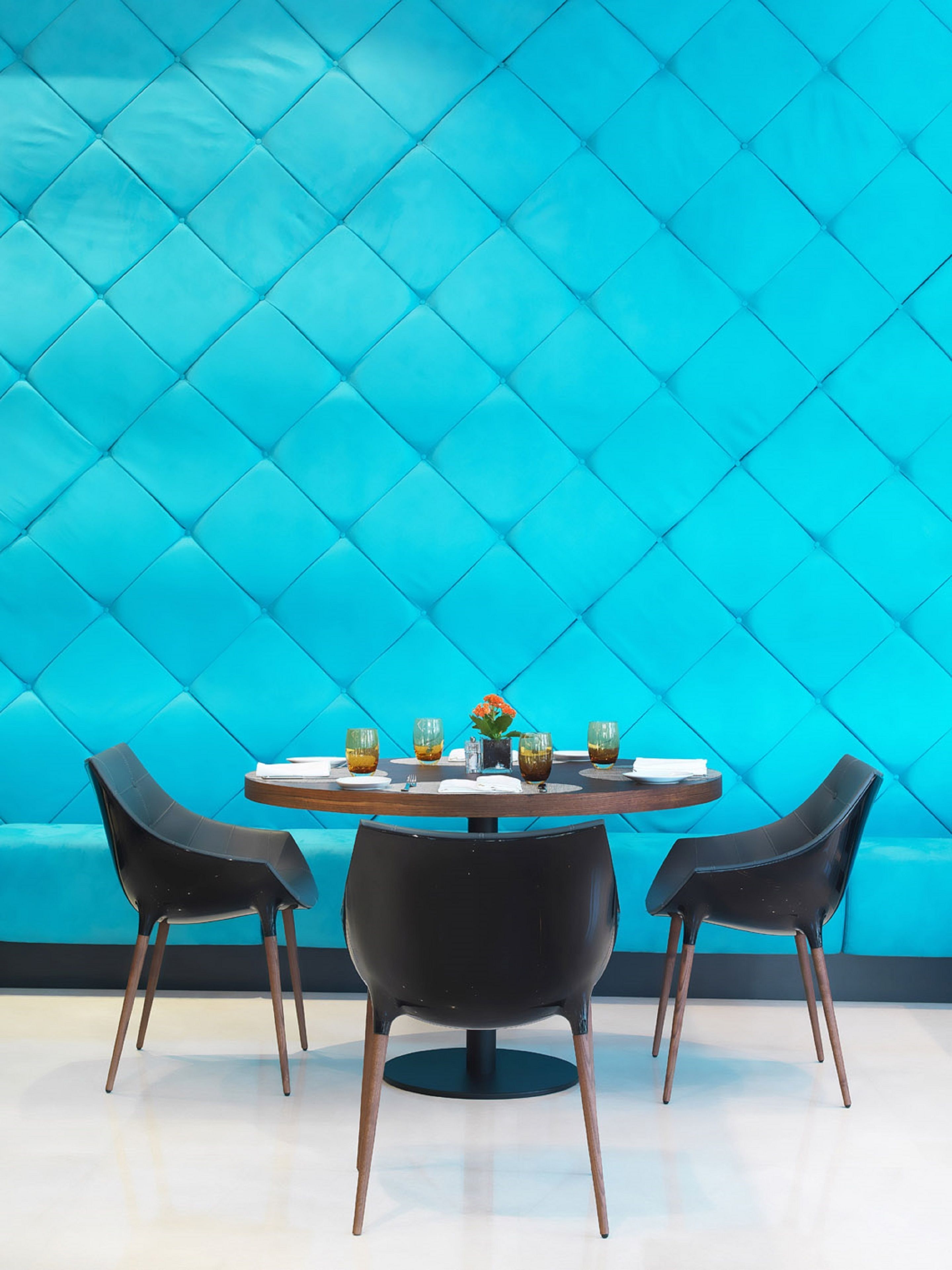 A round table with four chairs, a blue cushioned wall, and a small vase with flowers.