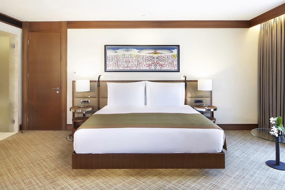 A hotel room with a large bed, two bedside tables with lamps, a painting on the wall, and a door to the left.
