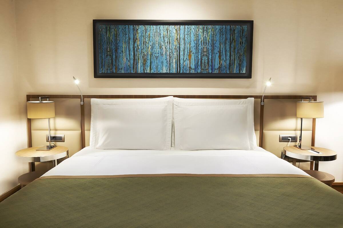 A hotel room with a large bed, two bedside tables with lamps, a framed artwork above the bed, and a green throw blanket on the bed.
