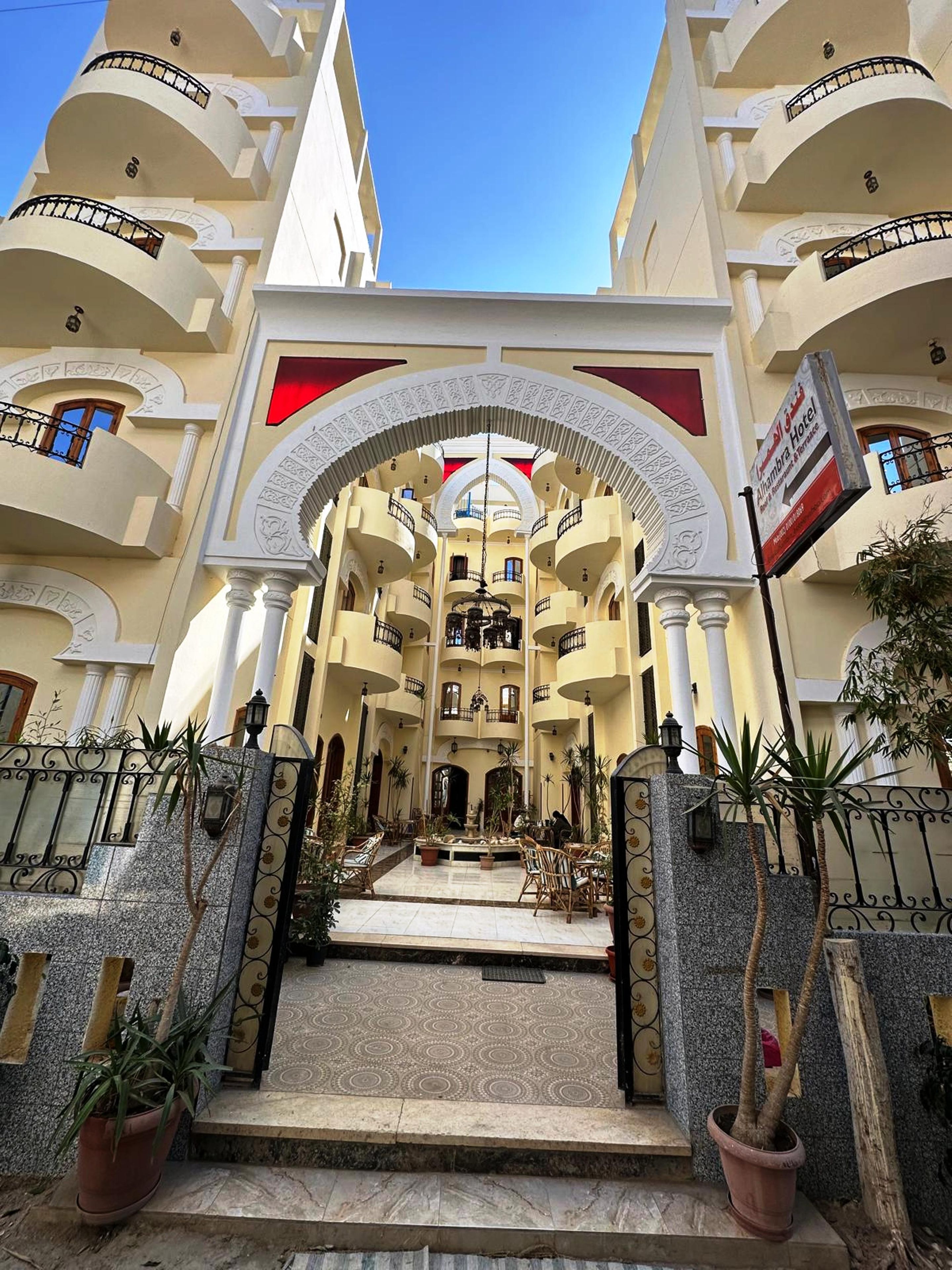 L’entrée d’un hôtel, avec un escalier central, de nombreux balcons et un drapeau indiquant le nom de l’hôtel.