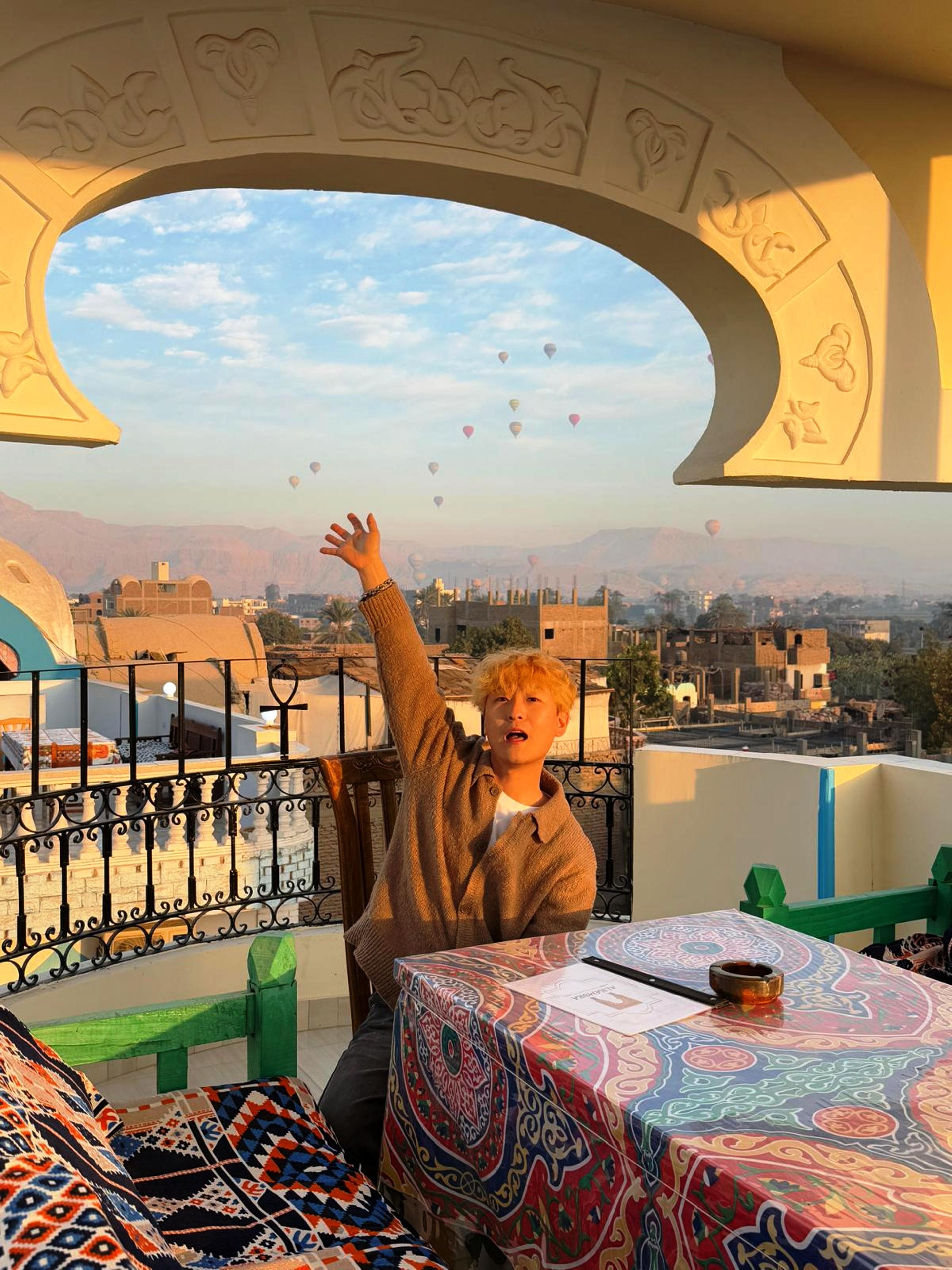 Una persona sentada a una mesa con mantel colorido, con vista a un paisaje urbano que incluye edificios y montañas, y una barandilla de balcón.
