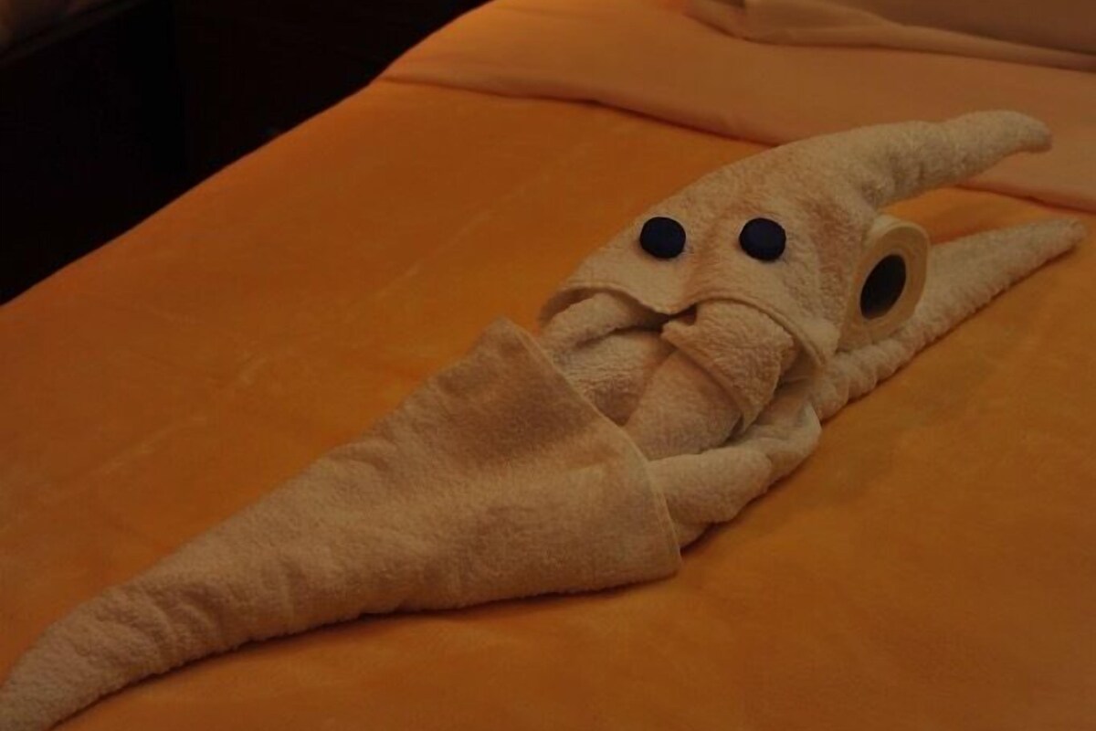 Una habitación de hotel con una cama, una mesita de noche con lámpara y una toalla doblada en forma de personaje.