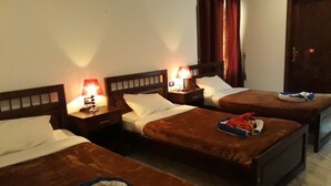 A hotel room with three beds, each with a wooden headboard and a nightstand with a lamp. There is a wooden door and a window with curtains.