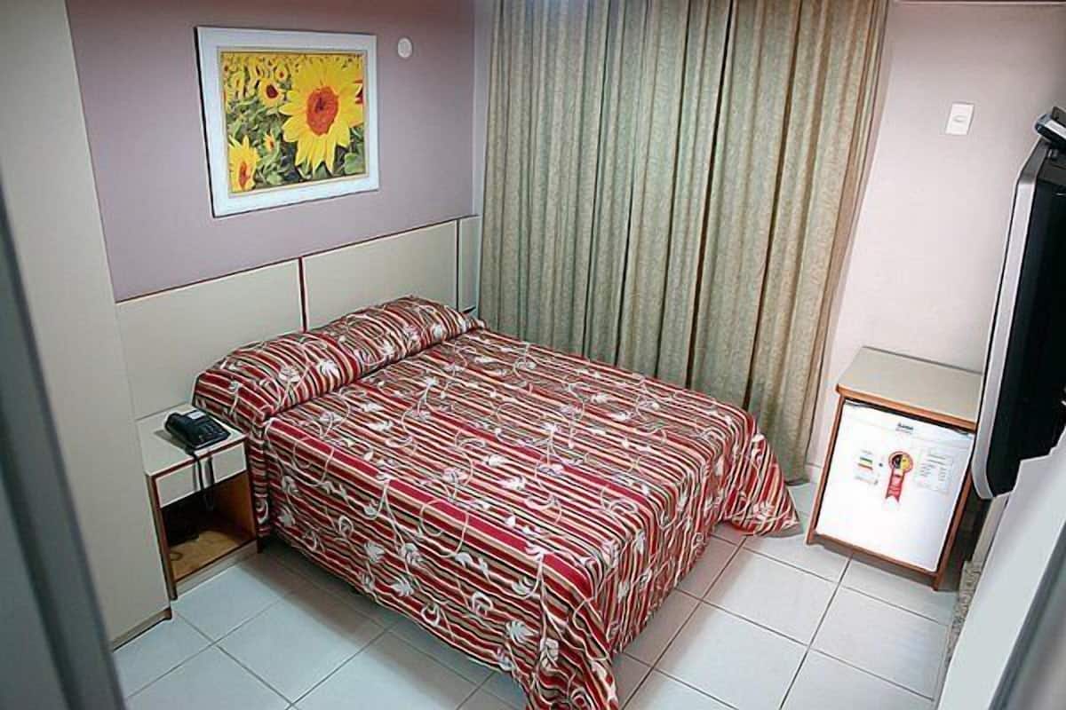 Un dormitorio pequeño y ordenado con una cama, una mesita de noche, un cuadro enmarcado en la pared y una pequeña mesa con una pantalla.