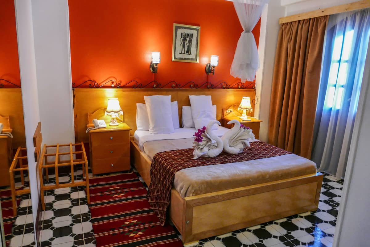 Habitación de hotel con una cama grande, dos mesitas de noche con lámparas, un cabecero de madera, un tapete con estampado rojo y negro, y un cuadro en la pared.