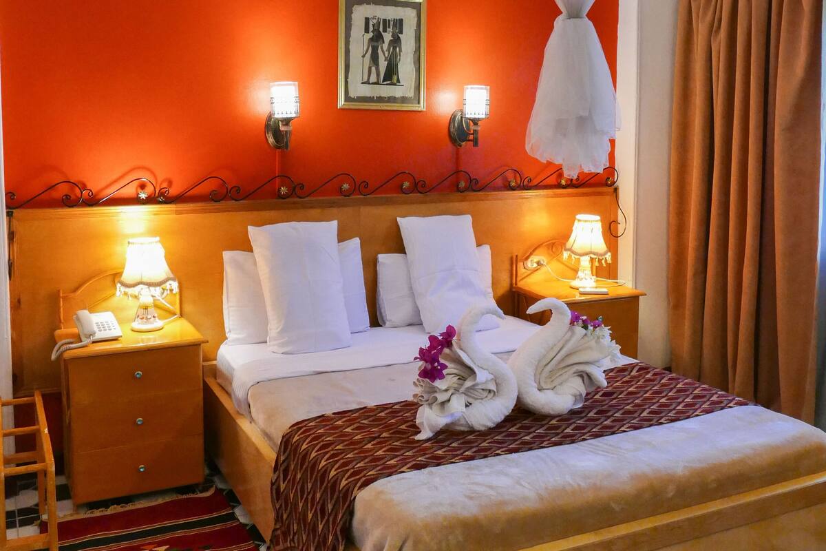 Habitación de hotel con cama, mesitas de noche, alfombra estampada en rojo y negro, mosquitero y un cuadro en la pared.