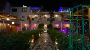 Ein Nachtblick auf einen Hotelinnenhof mit einem mosaikgepflasterten Weg, der zum Eingang führt, umgeben von Grünpflanzen und dekorativer Beleuchtung.