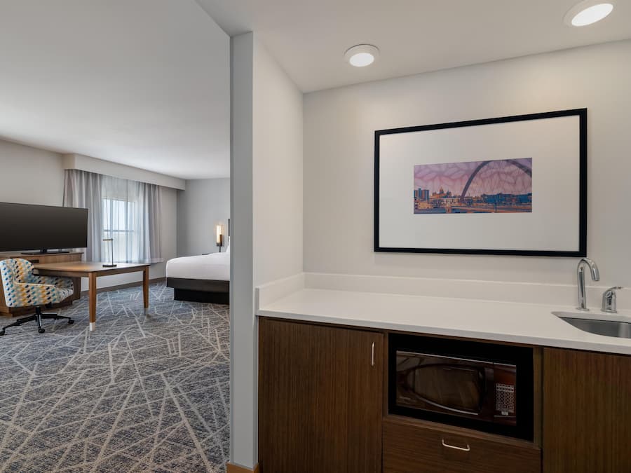 A hotel room with a bed, a desk with a chair, a television, a framed picture on the wall, a sink, and a microwave.